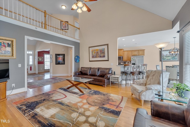 living room with baseboards, high vaulted ceiling, ceiling fan with notable chandelier, and light wood-style floors