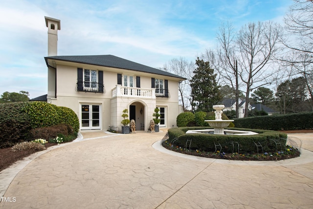 view of front of property with concrete driveway, stucco siding, french doors, a chimney, and a balcony