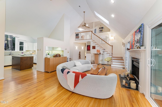 living room featuring a fireplace with raised hearth, high vaulted ceiling, a skylight, light wood-style floors, and stairway