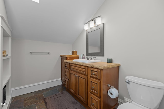 bathroom featuring toilet, vanity, baseboards, vaulted ceiling, and stone tile flooring