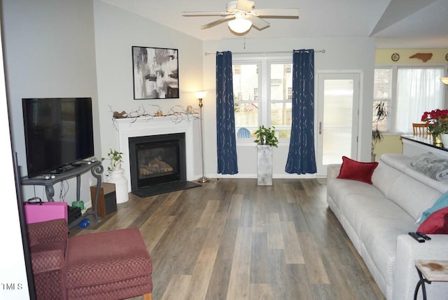 living room featuring wood finished floors, a ceiling fan, baseboards, vaulted ceiling, and a glass covered fireplace