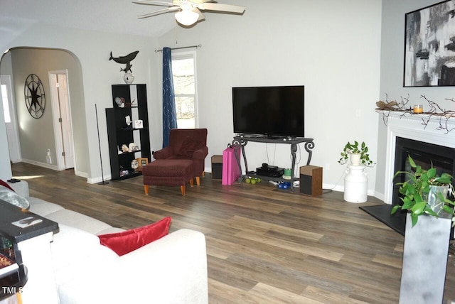 living room with arched walkways, a fireplace with raised hearth, a ceiling fan, wood finished floors, and vaulted ceiling