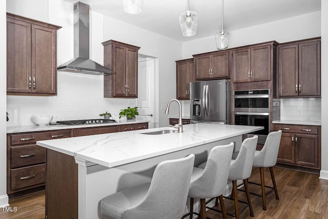 kitchen featuring dark wood finished floors, an island with sink, a sink, appliances with stainless steel finishes, and wall chimney exhaust hood