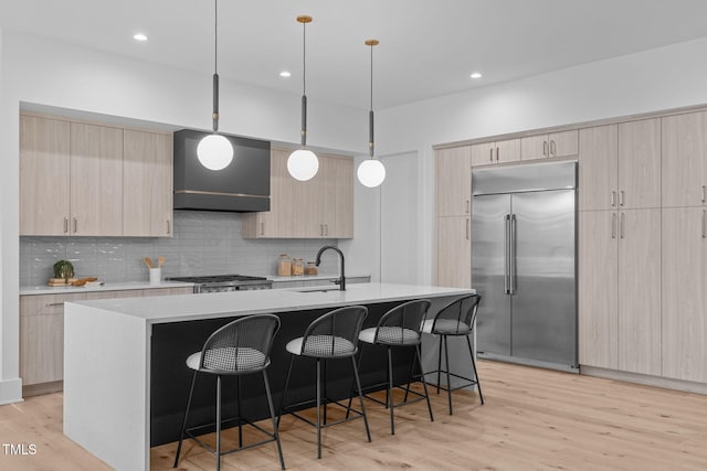 kitchen featuring built in refrigerator, light brown cabinets, hanging light fixtures, and an island with sink