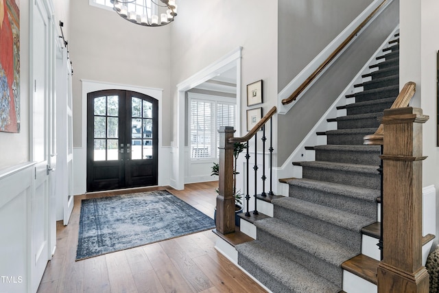 foyer entrance featuring a towering ceiling, a wainscoted wall, wood-type flooring, french doors, and a chandelier