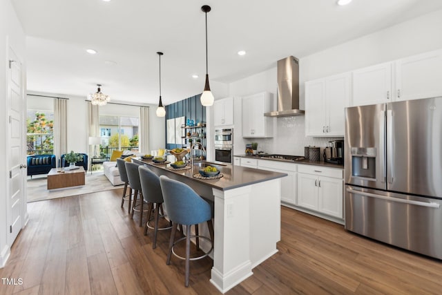 kitchen with dark countertops, appliances with stainless steel finishes, white cabinets, an island with sink, and wall chimney exhaust hood