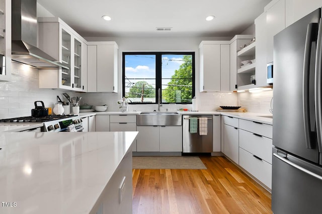 kitchen with stainless steel appliances, light countertops, a sink, wall chimney range hood, and light wood-type flooring