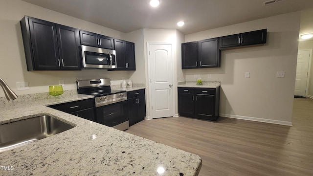 kitchen featuring dark cabinets, a sink, appliances with stainless steel finishes, light stone countertops, and light wood finished floors