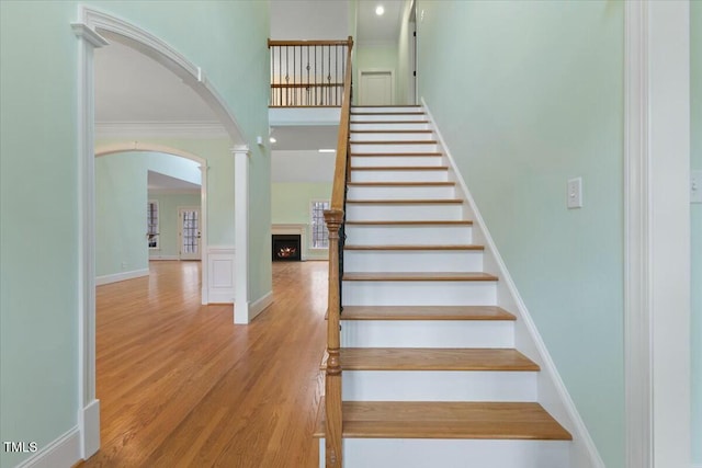 stairs with arched walkways, crown molding, a towering ceiling, wood finished floors, and a warm lit fireplace