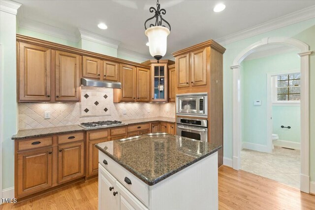 kitchen with arched walkways, stainless steel appliances, brown cabinetry, glass insert cabinets, and under cabinet range hood