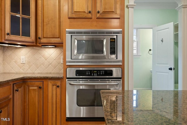 kitchen featuring brown cabinets, tasteful backsplash, appliances with stainless steel finishes, glass insert cabinets, and dark stone counters