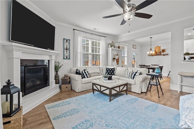 living area featuring visible vents, a glass covered fireplace, wood finished floors, crown molding, and ceiling fan with notable chandelier