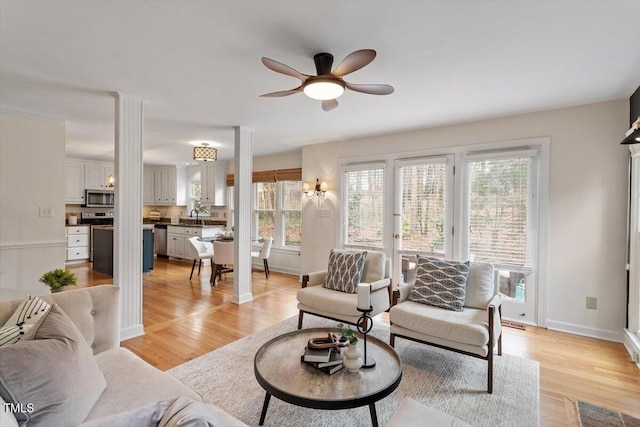 living area with light wood-type flooring, baseboards, a ceiling fan, and decorative columns