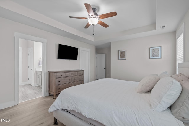 bedroom with a tray ceiling, visible vents, light wood-style floors, ensuite bath, and baseboards