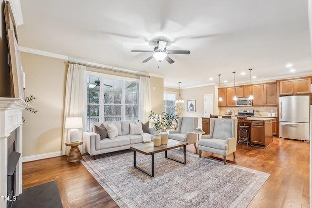 living room with ceiling fan with notable chandelier, a fireplace with flush hearth, light wood-style floors, baseboards, and crown molding