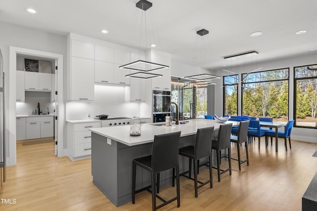 kitchen featuring light wood-type flooring, a center island with sink, a sink, white cabinets, and light countertops