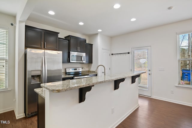 kitchen featuring stainless steel appliances, a breakfast bar, a center island with sink, and light stone countertops