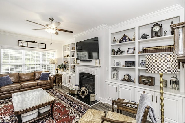 living area featuring built in features, ceiling fan, ornamental molding, a fireplace with raised hearth, and dark wood-style floors