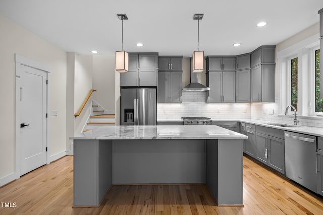 kitchen featuring a kitchen island, appliances with stainless steel finishes, gray cabinets, wall chimney range hood, and a sink