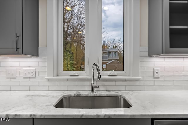 kitchen featuring gray cabinetry, a sink, light stone countertops, tasteful backsplash, and glass insert cabinets