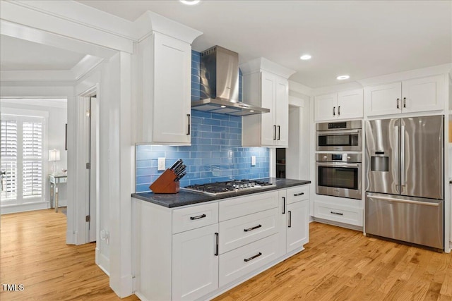 kitchen with white cabinets, appliances with stainless steel finishes, wall chimney exhaust hood, light wood finished floors, and dark countertops