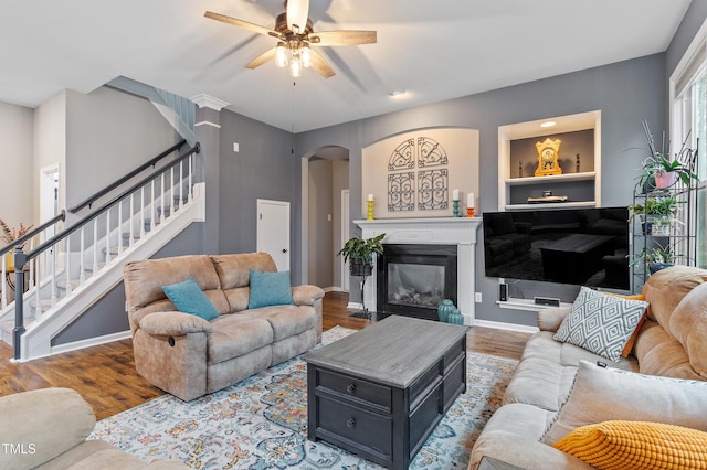 living room featuring a ceiling fan, a glass covered fireplace, wood finished floors, arched walkways, and stairs