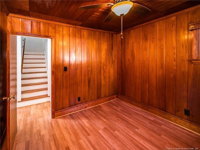 empty room with ceiling fan, stairway, light wood-style flooring, and wooden walls