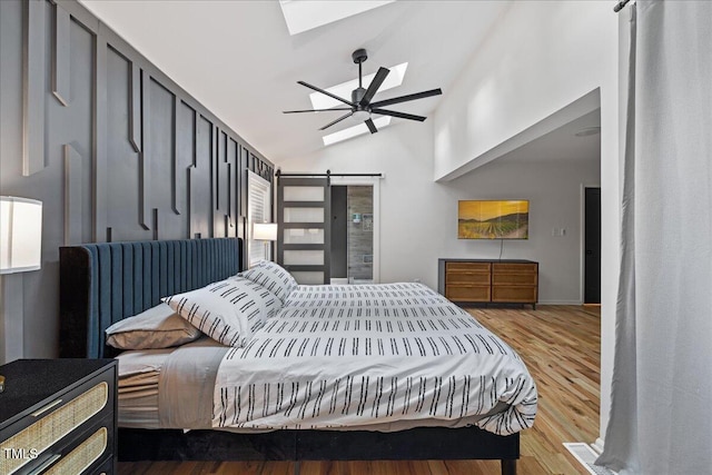 bedroom featuring a skylight, a barn door, a ceiling fan, light wood-style flooring, and high vaulted ceiling