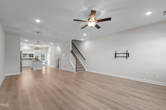 unfurnished living room featuring recessed lighting, ceiling fan with notable chandelier, baseboards, stairway, and light wood-type flooring