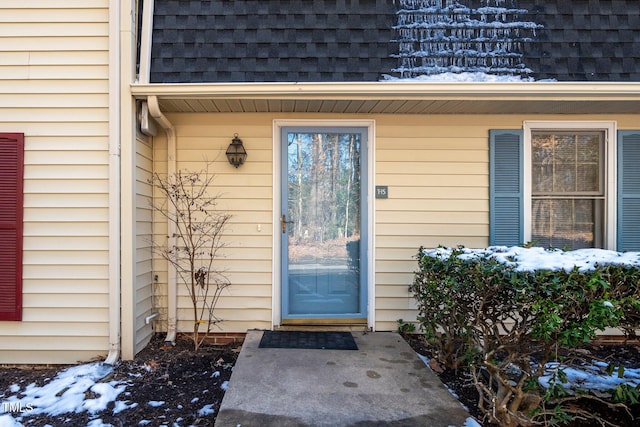 snow covered property entrance featuring a shingled roof and mansard roof