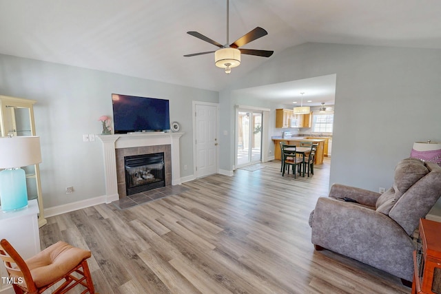 living area featuring a ceiling fan, baseboards, vaulted ceiling, light wood-type flooring, and a tiled fireplace