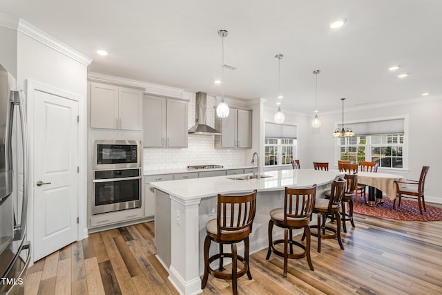 kitchen with light countertops, appliances with stainless steel finishes, a sink, wall chimney range hood, and an island with sink