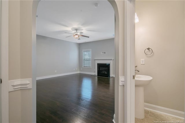 unfurnished living room with arched walkways, dark wood-style flooring, a fireplace with flush hearth, ceiling fan, and baseboards