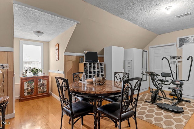 dining room with visible vents, vaulted ceiling, wainscoting, light wood-style flooring, and a textured ceiling