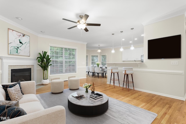 living area featuring ceiling fan, light wood-style flooring, a tile fireplace, ornamental molding, and baseboards