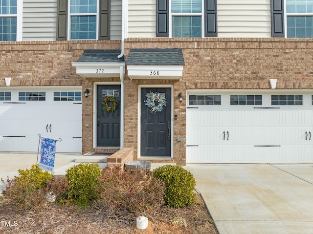 doorway to property featuring a garage, brick siding, roof with shingles, and concrete driveway