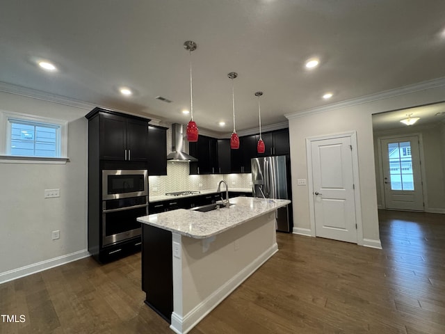 kitchen with a sink, hanging light fixtures, appliances with stainless steel finishes, wall chimney exhaust hood, and an island with sink