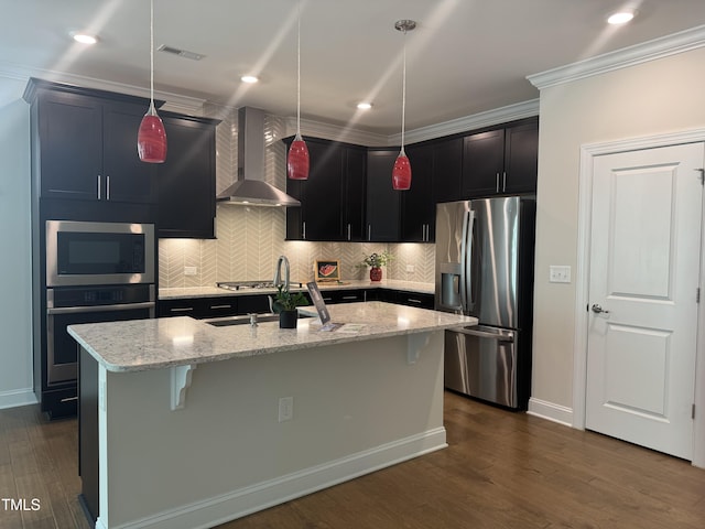 kitchen with wall chimney exhaust hood, appliances with stainless steel finishes, decorative light fixtures, light stone countertops, and a kitchen island with sink