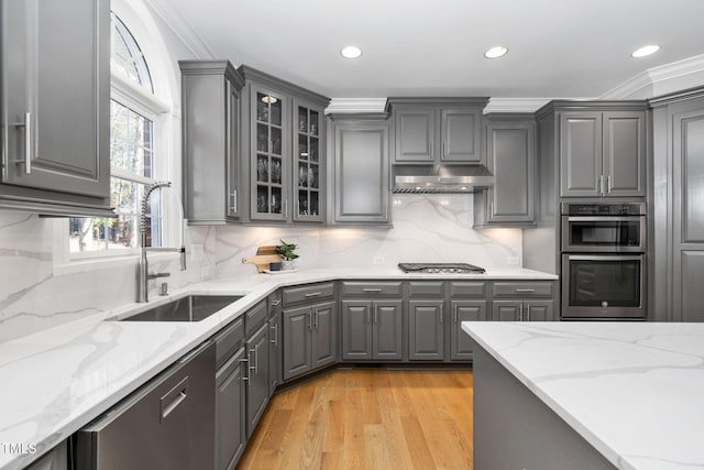 kitchen featuring light stone counters, gray cabinetry, glass insert cabinets, a sink, and under cabinet range hood