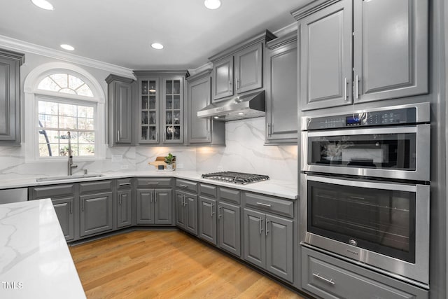 kitchen with light stone counters, appliances with stainless steel finishes, glass insert cabinets, a sink, and under cabinet range hood