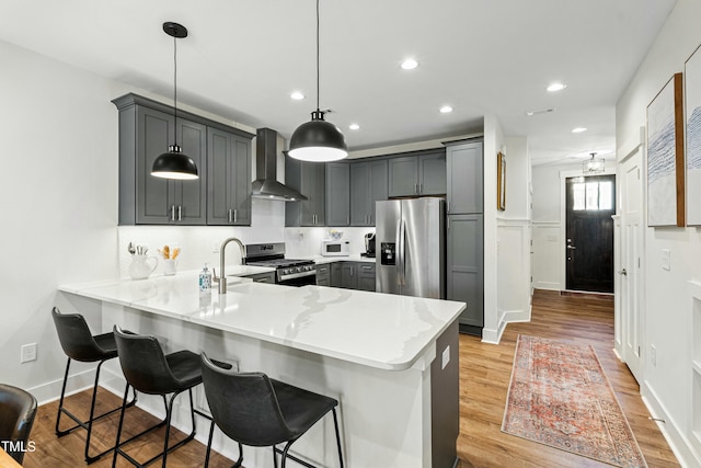 kitchen with a breakfast bar area, a peninsula, hanging light fixtures, appliances with stainless steel finishes, and wall chimney exhaust hood