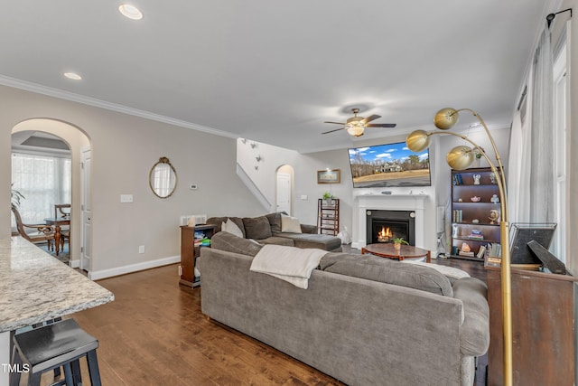 living room with arched walkways, dark wood-style flooring, crown molding, and a lit fireplace