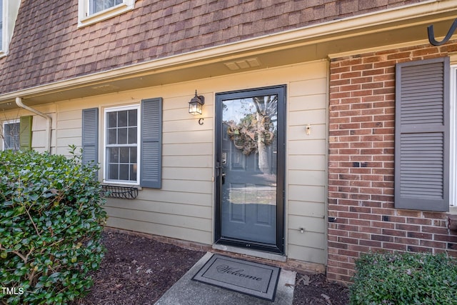 property entrance featuring brick siding, roof with shingles, and mansard roof