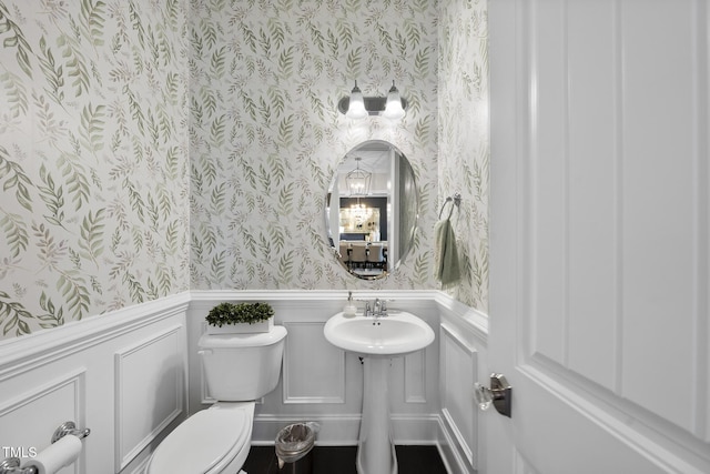 half bathroom featuring toilet, wallpapered walls, a decorative wall, and wainscoting