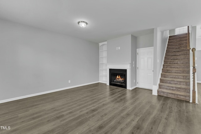 unfurnished living room with a lit fireplace, stairway, dark wood finished floors, and baseboards