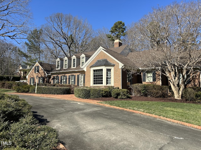 new england style home featuring driveway, brick siding, a chimney, roof with shingles, and a front yard