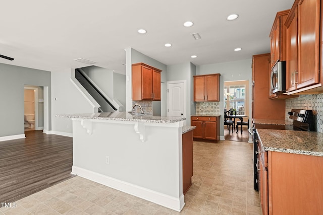 4508 Brintons Cottage St, Raleigh, NC 27616 - kitchen with appliances with stainless steel finishes, a peninsula, a breakfast bar, brown cabinetry, and light stone countertops