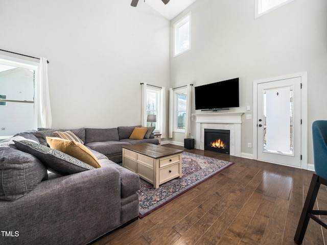 living room featuring baseboards, a towering ceiling, ceiling fan, dark wood-style flooring, and a lit fireplace