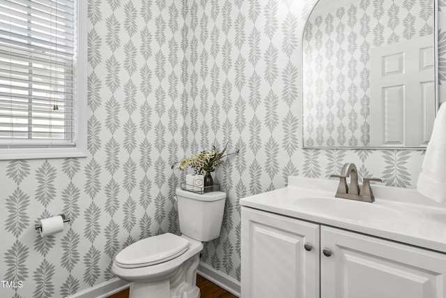 half bathroom with wallpapered walls, baseboards, toilet, wood finished floors, and vanity