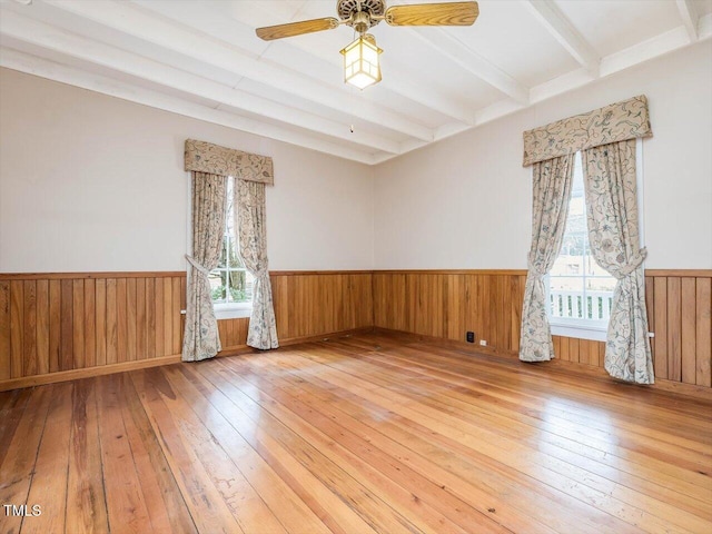 empty room featuring a wainscoted wall, wood-type flooring, a wealth of natural light, and beamed ceiling